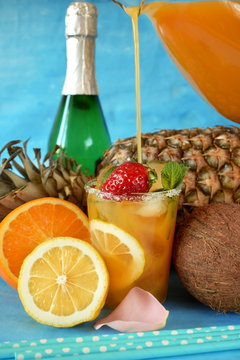 Glass with ice is being filled up with an orange drink surrounded by fruits. Champagne bottle in the background