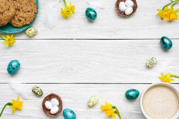 frame made of easter eggs, cup of cappuccino, spring flowers and biscuits on white wooden background. easter composition