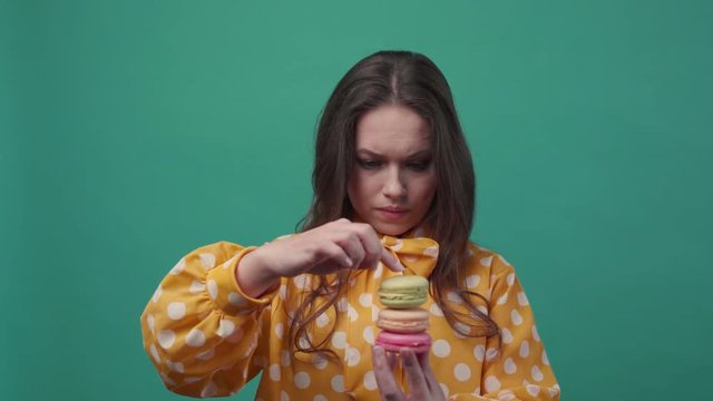Pretty young woman puts three macarons one on top of another, wants to eat it all at the same time on a colorful background. Having fun, delicious dessert. Slow motion, female portrait