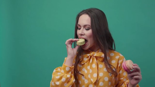 Gorgeous young woman bites the macaroon and holds two more in her hand, looks straight to camera, feels tasty. Gourmet, sweet tooth. Slow motion, close up view, female portrait