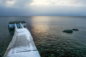 Wooden boat pier covered with ice and snow with icicles at the end, with sun shining on sea in background.