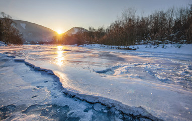 River Bela completely frozen during extreme cold, early morning sun rising behind mountains reflected in ice layer with snow patches. Liptovsky Hradok, Slovakia