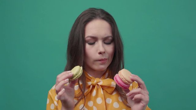 Attractive brunette woman on a colorful background, licks her lips, looks at cookies choosing between one macaron and two macarons, sighs, and eats the green one, closes her eyes with pleasure