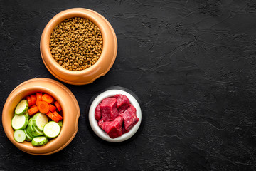 pet care with dry food, vegetables and raw meat for dog or cat in plastic bowl on black background top view space for text