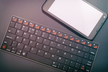 Wireless keyboard and smartphone with blank screen on dark background. Toned