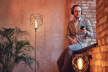 Portrait of a stylish man in a flannel suit and glasses listening music with the phone and headphones sitting on a window sill in a room with loft interior.