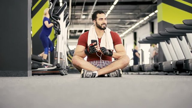 Handsome muscular man wears red t-shirt with towel on shoulders sitting in the gym using smartphone, workout rest pause