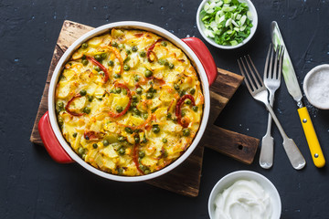 Potatoes and vegetables tortilla on a dark background, top view. Potatoes, green beans, bell peppers, green peas, cheese, eggs casserole in a baking dish -  delicious breakfast or snack - obrazy, fototapety, plakaty