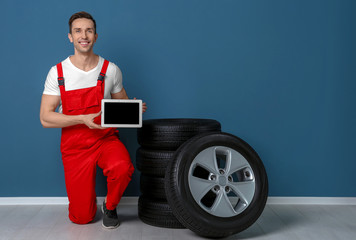 Young mechanic in uniform with tablet PC and car tires near color wall