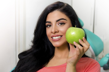 Gorgeous young woman holding green apple next to perfect white teeth , while sitting in dentist chair 