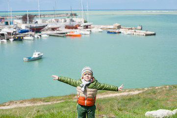 Fototapeta na wymiar dreamy little kid in jacket and hat stands on green hill spreading his hands like wings on background of sea bay with boats in early spring