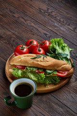 Fresh sandwich with lettuce, tomatoes, cheese on wooden plate, cup of coffee on wooden background, selective focus