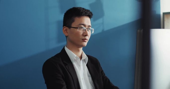 Portrait young chinese man with glasses uses computer to look at the screen in office business internet face technology businessman laptop male sitting job attractive interior handsome slow motion