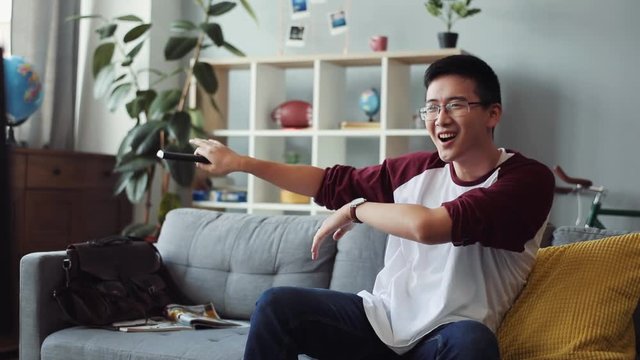 attractive young chinese man with glasses sits on the couch dancing and turns on the TV at home looking house technology male business happy leisure video sofa portrait close up slow motion