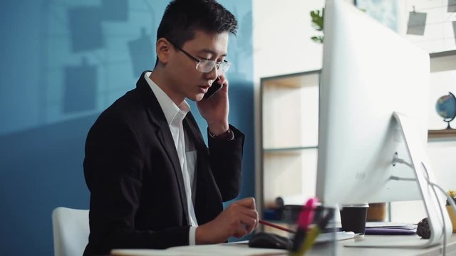 Man seriuos young chinese with glasses uses computer speak on phone write documents office business internet face technology businessman laptop male sit job attractive interior handsome slow motion