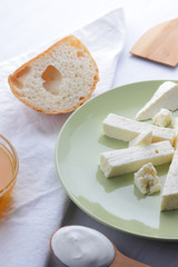 Cheese, honey and sour cream, a piece of bread, feta in a green plate, soft cheese on a white background, a wooden spoon with sour cream, French breakfast, blue napkin, honey in glassware, art