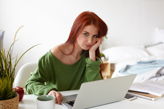 Picture of beautiful casually dressed European student girl with red hair having bored expression while doing research project at home, sitting at workplace with laptop, phone and textbooks on desk
