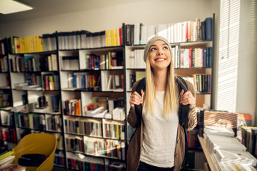 A young please female student is happily looking through the window in front of library bookshelf while holding her backpack sleeves.