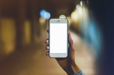 hipster using in hands clean gadget mobile phone closeup, blogger pointing finger on blank screen smartphone on background bokeh light in night atmospheric city, mockup online wifi internet concept