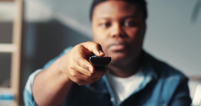 Close up face serious african american man watching tv sitting on the couch holds the remote changing channel commercials flipping guy handsome happy home interior technology portrait slow motion