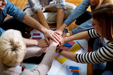 Close up view of the young  motivated team of business people putting hands together between them while sitting on the office floor.