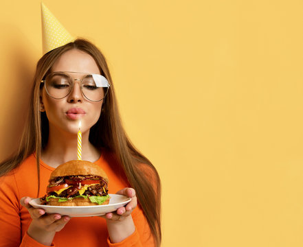 Woman with tasty big beef burger barbeque sandwich for birthday party with lit candle on pastel yellow