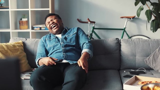Portrait rude african american man watching tv sit on couch laughs smile happy changing channel commercials control flipping guy handsome cute home interior technology portrait slow motion
