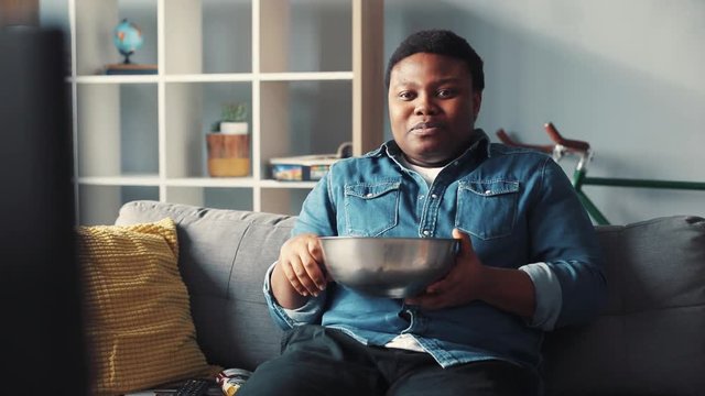 Young african american man watching tv sit on couch eat chips smile laughs happy changing channel commercials control flipping guy handsome cute home interior technology portrait slow motion