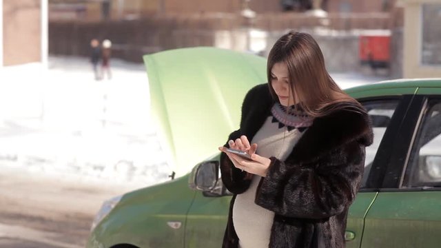 In winter, a pregnant girl, standing near a faulty car with an open hood, trying to call a tow truck with a mobile phone