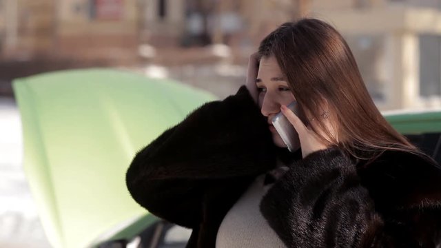 In winter, a pregnant girl, standing near a faulty car with an open hood, trying to call a tow truck with a mobile phone