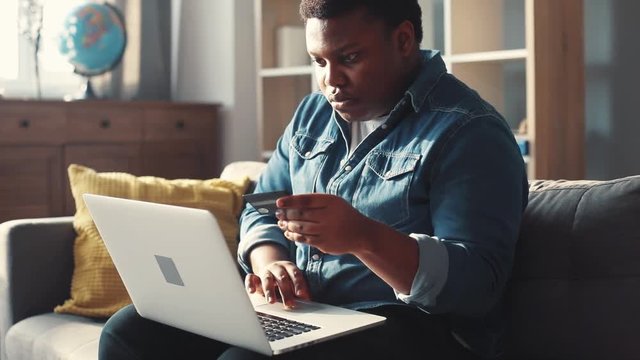 Serious young african american man use laptop and credit card sit on sofa in home online relax work cell phone shoping sunset guy close up mobile handsome portrait slow motion rude attractive