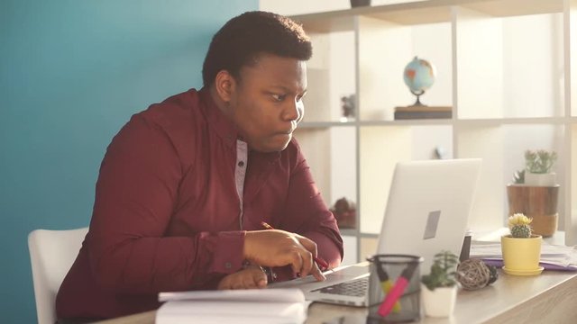 Thinking young african american man writing and working on laptop in office sunlight business person internet face businessman freelance looking portrait serious work slow motion black brainstorming