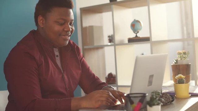 Happy young african american man working on laptop typing in office smiling sunlight business person internet face businessman freelance looking portrait work slow motion black brainstorming