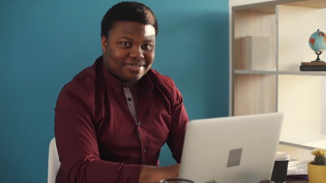 Close up young african american man working on laptop look at camera smile in office happy sunlight business internet face businessman freelance looking portrait work slow motion black brainstorming