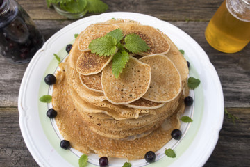 Large and small pancakes are located in a plate on an old wooden table decorated with berries and mint leaves.