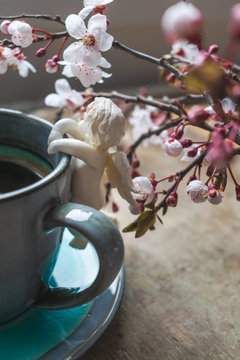 View of a white angel figurine hanging on a vintage cup of coffee on a rustic, old wooden table and blooming tree branches in a vase. Hello spring or Happy Easter background or concept. Toned image
