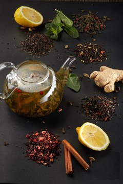 Image for a menu or poster with a choice of tea or ingredients. The image has a transparent teapot with tea inside on a black background and sprinkled with tea, lemon, ginger, mint, cinnamon