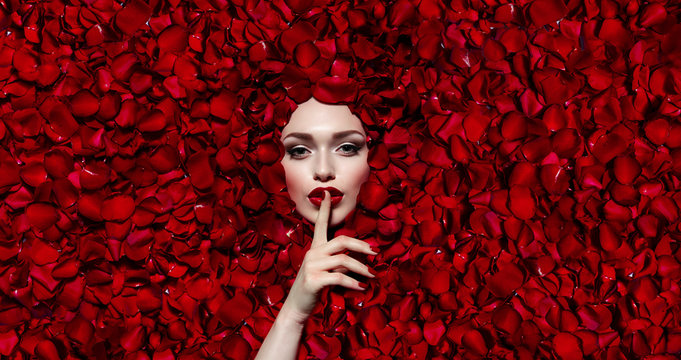 Portrait of a beautiful young girl in red rose petals. Red lips, red fingernails. The girl makes a gesture with her hand.Fashion, beauty, make-up, cosmetics, hairstyle, beauty salon, boutique, discoun