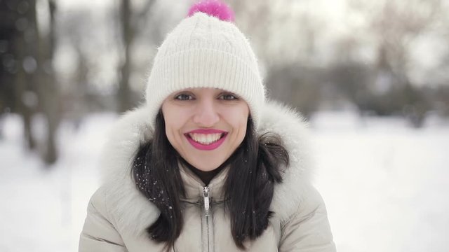 Closeup Brunette hair young women portrait with pink lips in beige down coat and hat throws up snow and smiling in snowy central park, snowfall. Slow motion.