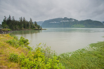 Der Skeena River am Yellowhead Hwy, Highway nach Port Rupert, tr&uuml;bes Wetter, Regen, Wolkenverhangene Berge im Hintergrund, bewaldete Insel links im Fluss. Vorherschende Farbe grau gr&uuml;n.