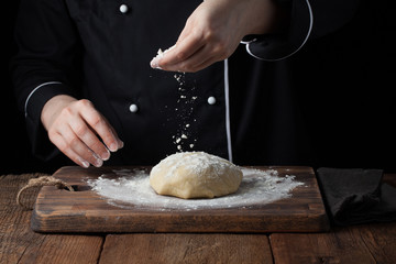 Chef hands pouring flour powder on raw dough using sieve on a black background, Cooking process