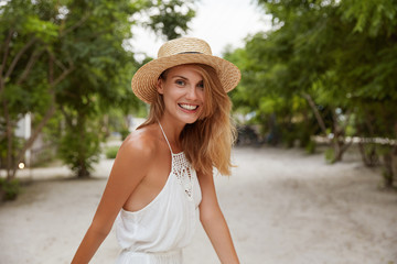 Pretty smiling young female with glad expression, wears fashionable white dress and hat, strolls outdoor, has tanned healthy skin, enjoys summer vacations. People happiness and lifestyle concept