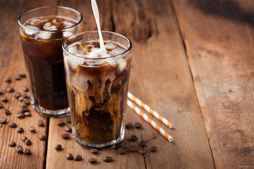 Ice coffee in a tall glass with cream poured over and coffee beans on a old rustic wooden table. Cold summer drink on a dark wooden background with copy space