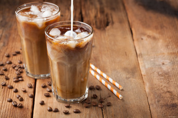 Ice coffee in a tall glass with cream poured over and coffee beans on a old rustic wooden table. Cold summer drink on a dark wooden background with copy space