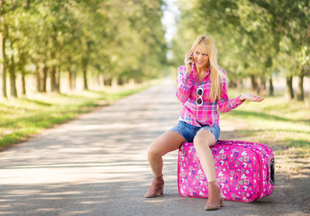 Upset young blonde women sitting on suitcase and talking on smartphone along the road. Beautiful female traveler going on vacation