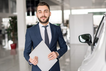 Waist up portrait of handsome car salesman looking at camera  and smiling holding clipboard standing in luxury dealership showroom