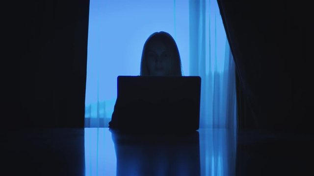 Business woman working on laptop computer sitting back to window in dark room. Young woman using notebook for overtime work in night home studio