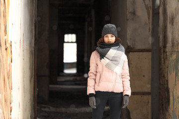 A young cute white girl in a hat warm scarf and gloves is standing in the corridor of a ruined house in winter. Destroyed building.