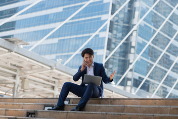 Young Asian businessman talking on mobile phone with a serious face and Working at laptop Sitting in a city Bangkok Thailand. Concept of communication.