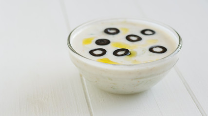 Tzatziki sauce bowl decorated with olives and oil on a white wooden table.